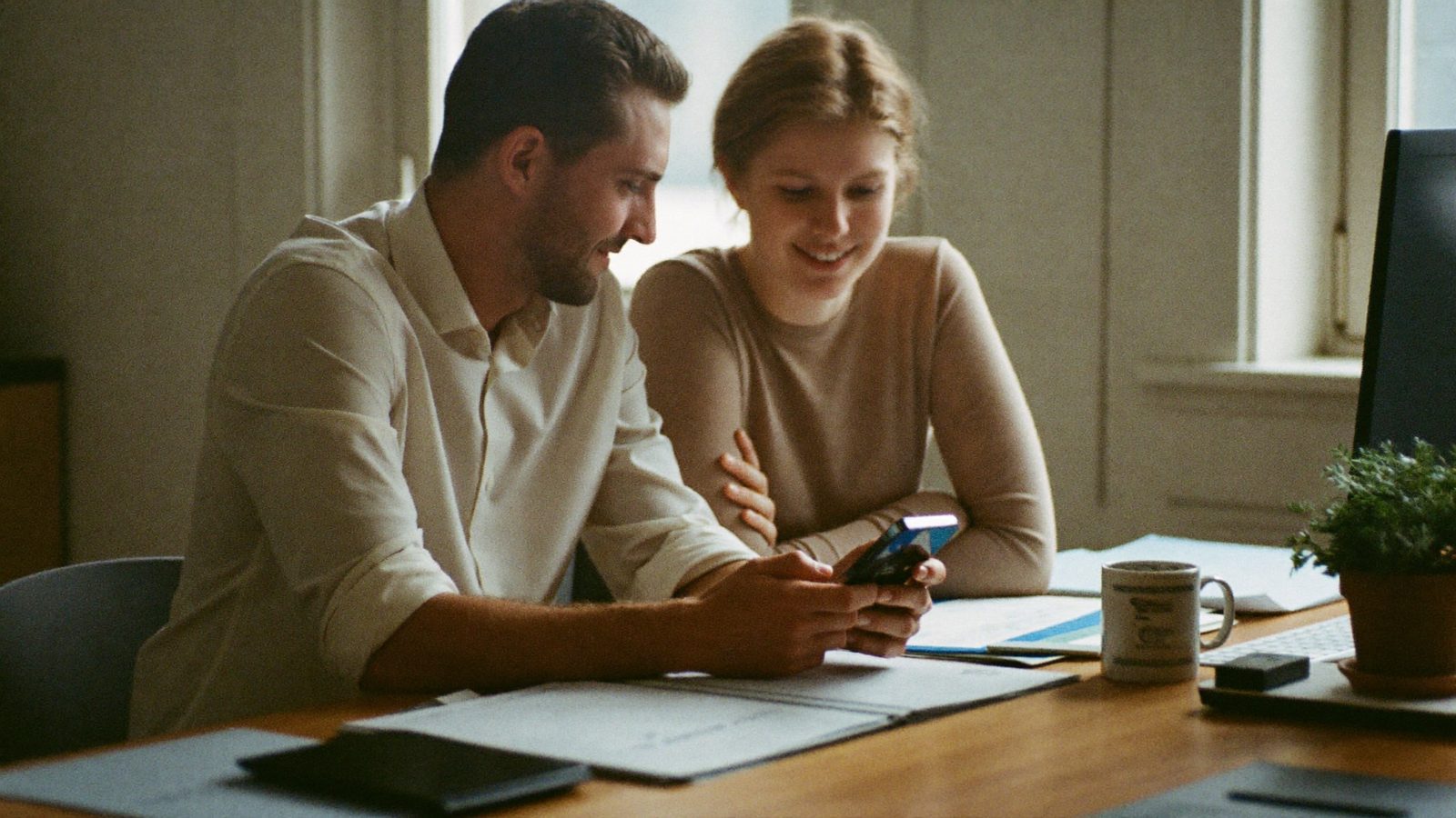 Zwei Menschen am Schreibtisch schauen zufrieden auf ein Smartphone mit einem Chatverlauf